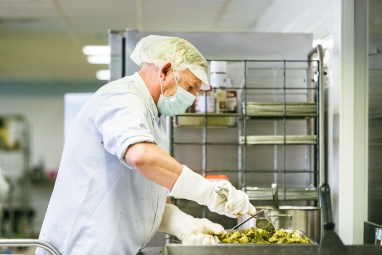 Une personne portant un filet à cheveux, un masque facial, des gants chirurgicaux et un tablier bleu clair prépare de la nourriture dans une cuisine. Elle prend de la nourriture dans un plateau. Derrière elle, il y a un support en métal avec des étagères contenant divers contenants et fournitures. Le décor de la cuisine propre et organisé reflète le dévouement des Héros Méconnus du CH d'Angoulême.