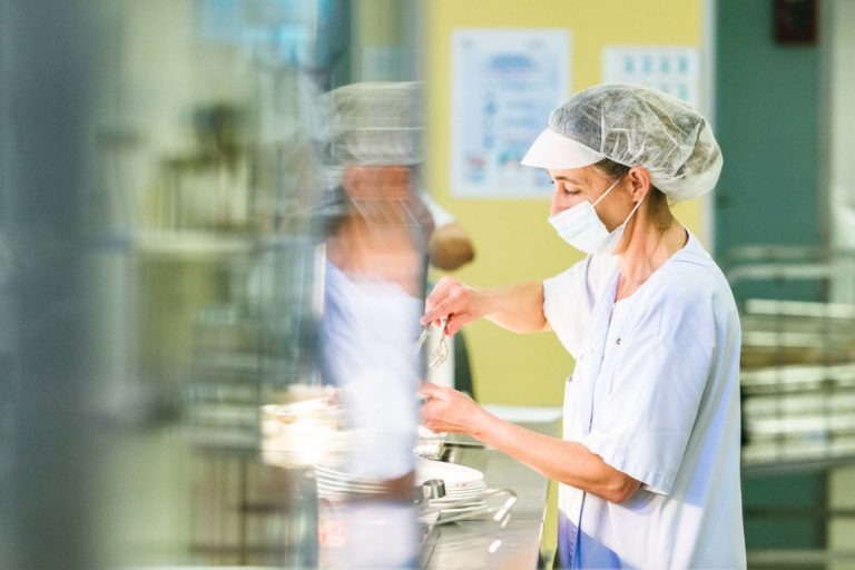 Une travailleuse de la santé en uniforme blanc, filet à cheveux et masque facial prépare un repas sur un comptoir en métal. Plusieurs assiettes sont alignées devant elle. L'arrière-plan montre des murs jaunes et verts et un reflet flou de la travailleuse sur la surface brillante près d'elle – vraiment l'un des Héros Méconnus du CH d'Angoulême.