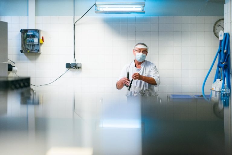 Une personne portant une blouse blanche, un masque et un filet à cheveux se tient debout dans une pièce aux murs carrelés, peut-être un laboratoire ou une cuisine. Elle tient un équipement en acier inoxydable et est éclairée par un plafonnier à économie d'énergie. Un point d'eau avec des tuyaux est visible sur le côté droit de l'image, emblématique des Héros Méconnus du CH d'Angoulême.