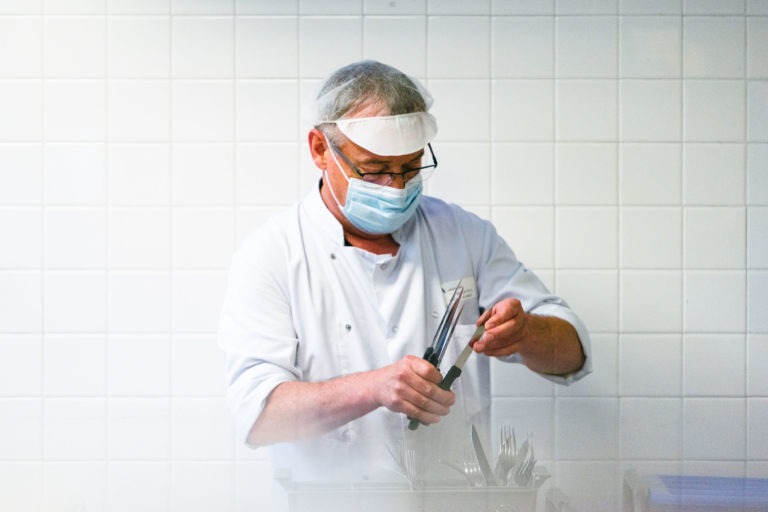 Un chef vêtu d'un uniforme blanc, d'une visière transparente et d'un masque chirurgical aiguise un couteau dans une cuisine aux murs carrelés de blanc. Il utilise un fusil à aiguiser pour aiguiser le couteau, en se concentrant intensément sur la tâche. Parmi les divers ustensiles posés sur le comptoir, il incarne l'un des Héros Méconnus du CH d'Angoulême.