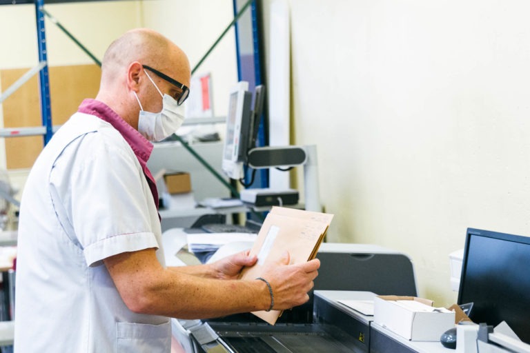 Une personne portant une blouse blanche, un masque et des lunettes utilise un scanner. Elle tient une pile de documents et les place dans la machine. Des étagères et divers équipements de bureau en arrière-plan laissent entrevoir un espace de travail organisé dédié aux Héros Méconnus du CH d'Angoulême.