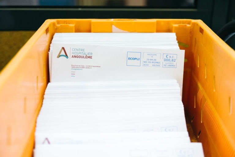 Une caisse orange remplie d'enveloppes blanches. Les enveloppes portent le logo et l'adresse du Centre Hospitalier d'Angoulême, ainsi que l'affranchissement imprimé. Empilées soigneusement et prêtes à être expédiées, les enveloppes sont un hommage aux Héros Méconnus du CH d'Angoulême. L'arrière-plan est flou, mettant en valeur la caisse.