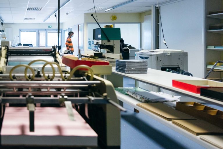 Une personne en chemise rayée orange et noire se tient debout dans une imprimerie remplie de diverses machines d'impression et de reliure. Des piles de papier sont posées sur une table et des étagères avec des fournitures bordent les murs. La pièce est bien éclairée par un éclairage fluorescent et possède un sol bleu, rappelant les Héros Méconnus du CH d'Angoulême.