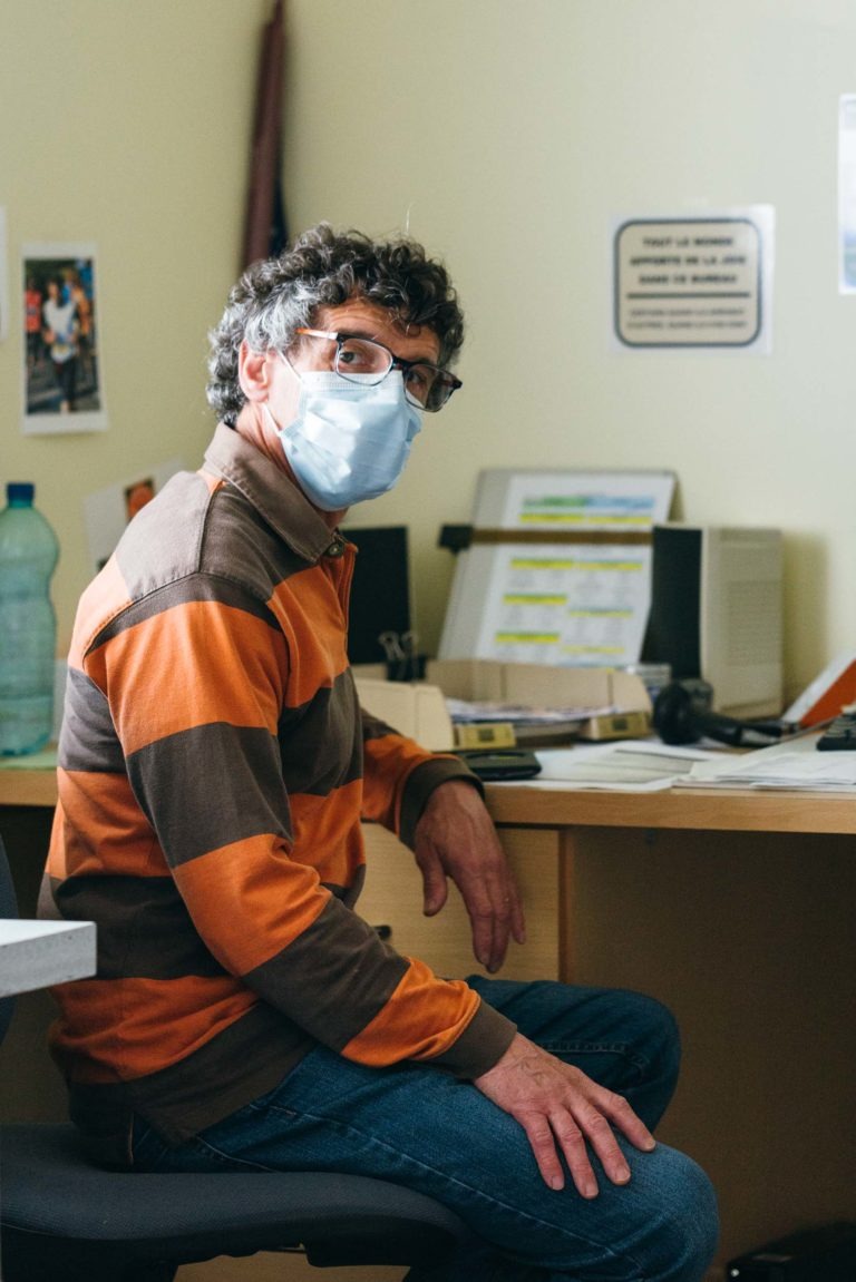 Un homme portant des lunettes et un masque bleu est assis sur une chaise de bureau, vêtu d'un pull rayé orange et marron. Son espace de travail, orné de l'emblème des Héros Méconnus du CH d'Angoulême, abrite un écran d'ordinateur, divers papiers et des fournitures de bureau. Des affiches et des avis décorent le mur beige derrière lui.