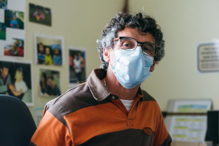 Un homme portant des lunettes et un masque jetable est assis à l'intérieur. Il a les cheveux gris bouclés et est vêtu d'une chemise rayée marron et orange. L'arrière-plan présente une variété de photos affichées sur le mur, l'une d'elles mettant en évidence les « Héros Méconnus du CH d'Angoulême », suggérant un cadre personnel ou professionnel.