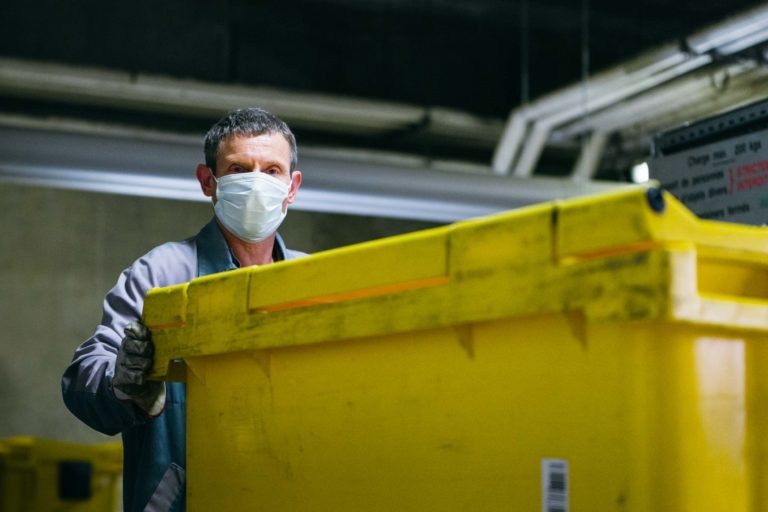 Une personne portant un masque et des gants se tient derrière une grande poubelle en plastique jaune dans un environnement industriel. L'individu, l'un des Héros Méconnus du CH d'Angoulême, tient la poubelle et semble la gérer ou la déplacer. L'arrière-plan comprend des équipements industriels et des tuyaux, indiquant un environnement de travail.