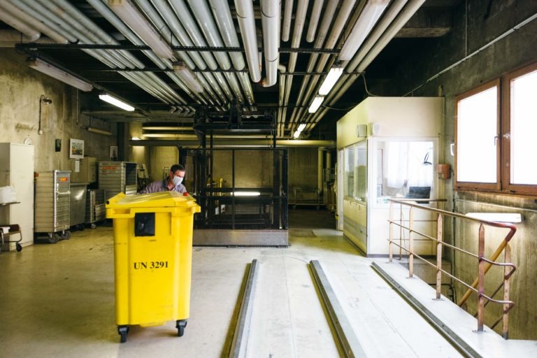 Une personne portant un masque pousse une grande poubelle jaune étiquetée « UN 3291 » dans un local industriel dont les tuyaux sont exposés au plafond. Avec de grandes fenêtres sur la droite et des balustrades métalliques, l'espace ressemble à une scène des « Héros méconnus du CH d'Angoulême ». Des étagères et une salle de contrôle fermée sont visibles en arrière-plan.