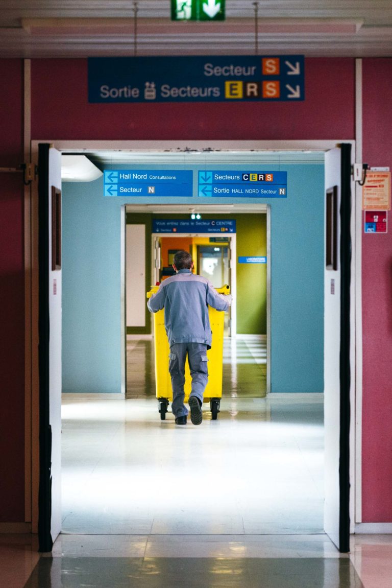 Un concierge en uniforme bleu pousse un grand chariot jaune dans un couloir d'hôpital très éclairé du CH d'Angoulême. Des panneaux de direction en français et en anglais indiquent les différents secteurs et sorties. Le sol propre et carrelé blanc brille sous les lumières, rendant hommage aux Héros Méconnus du CH d'Angoulême.