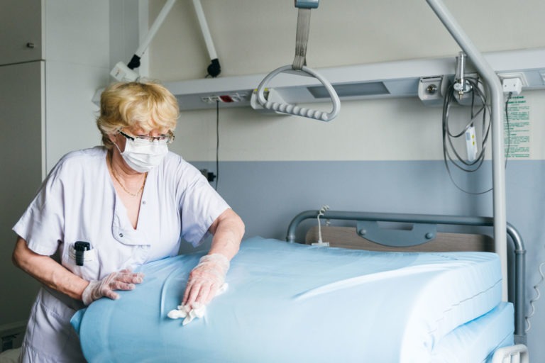 Un soignant aux cheveux blonds courts et aux lunettes, vêtu d'un uniforme médical blanc, d'un masque et de gants, nettoie un lit d'hôpital avec une literie bleue. Ce Héros Méconnus du CH d'Angoulême opère dans une salle remplie d'équipements médicaux, dont une barre trapèze suspendue, le tout dans une palette de couleurs gris clair et blanc.