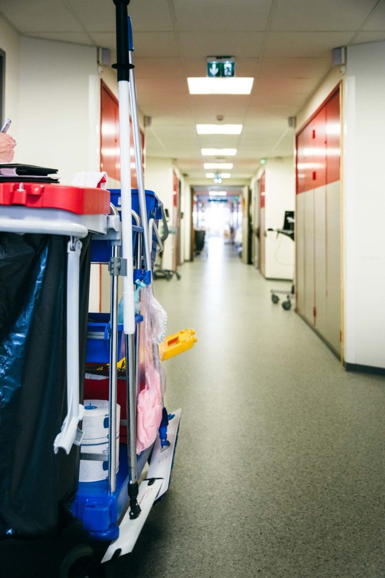 Un chariot de conciergerie rempli de produits de nettoyage, dont des serpillères et des sacs poubelles, se trouve dans un long couloir bien éclairé d'un bâtiment, peut-être un hôpital ou un bureau. Le sol gris, les murs rouges et beiges et les multiples portes fermées préparent le terrain pour que les Héros Méconnus du CH d'Angoulême maintiennent inlassablement la propreté.