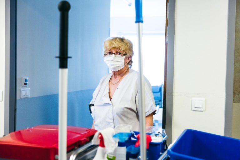 Une femme portant un uniforme médical blanc et un masque se tient dans une entrée, entourée de produits de nettoyage. Elle a les cheveux blonds courts et bouclés et des lunettes. Devant elle se trouve un chariot avec des plateaux rouges et bleus, un vaporisateur et divers outils de nettoyage. Elle représente les Héros Méconnus du CH d'Angoulême dans une salle lumineuse et partiellement éclairée.