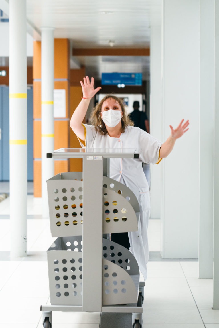 Un soignant portant un masque et un uniforme blanc se penche en avant de manière enjouée au-dessus d'un chariot d'hôpital dans un couloir lumineux et moderne. Les bras du soignant sont levés et les mains écartées, exprimant un geste joyeux et plein d'entrain, incarnant l'esprit des Héros Méconnus du CH d'Angoulême. Des piliers et des panneaux en arrière-plan indiquent un cadre hospitalier ou clinique.