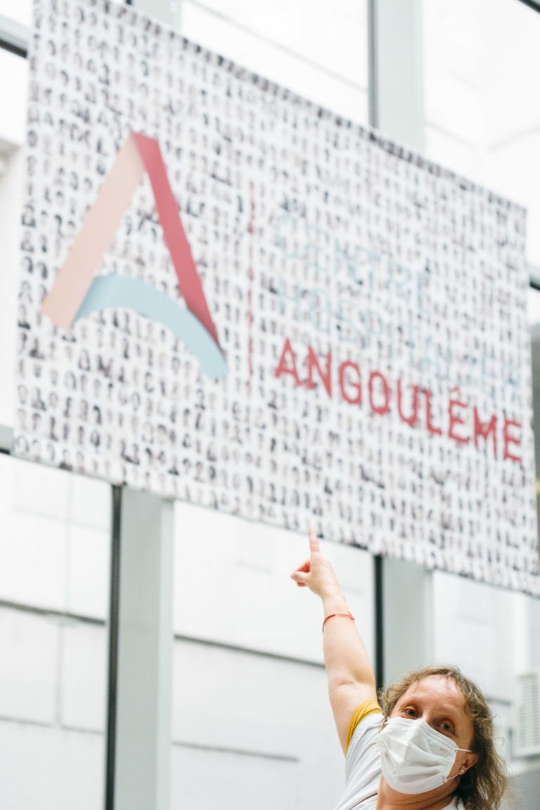 Une personne portant un masque blanc pointe vers le haut en direction d'un grand panneau. Le panneau comporte une lettre stylisée « A » en rouge et bleu, ainsi que le mot « ANGOULÊME » en lettres majuscules rouges. L'arrière-plan du panneau présente des vignettes de visages en noir et blanc, célébrant les Héros Méconnus du CH d'Angoulême à l'intérieur.