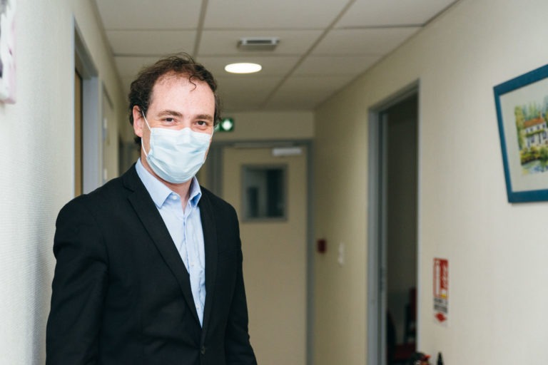 Un homme aux cheveux bouclés, portant un masque bleu, un blazer noir et une chemise bleue, se tient debout dans un couloir. Les murs clairs présentent un tableau représentant une maison sur la droite, avec des portes des deux côtés et un extincteur visible en arrière-plan. Il se tient parmi les Héros Méconnus du CH d'Angoulême.