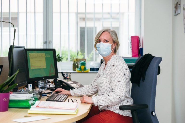 Une femme aux cheveux blonds courts, vêtue d'un chemisier blanc à petits motifs et d'un pantalon rouge, est assise à un bureau dans un bureau. Elle porte un masque chirurgical et travaille sur un ordinateur. Le bureau est encombré de papiers, clavier, souris, plante et autres fournitures de bureau ; elle incarne les Héros Méconnus du CH d'Angoulême. Le bureau dispose de grandes fenêtres avec