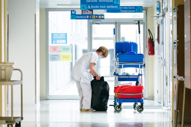 Un soignant en uniforme blanc et masque facial se penche pour ramasser un grand sac poubelle noir dans le couloir d'un hôpital. Un chariot de nettoyage avec des conteneurs bleus et rouges se trouve à proximité. Des panneaux suspendus et diverses affiches d'information sont visibles en arrière-plan, mettant en avant les Héros Méconnus du CH d'Angoulême.