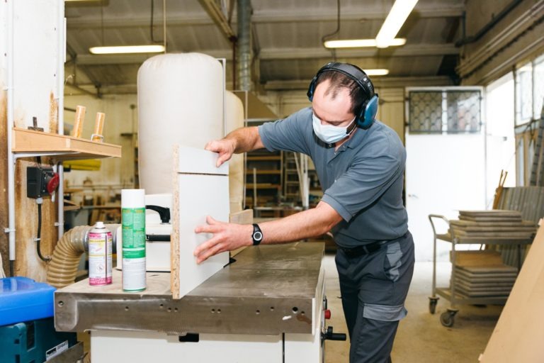 Une personne portant une chemise grise, des écouteurs de protection et un masque facial travaille dans un atelier bien éclairé. Elle utilise une scie circulaire pour couper un morceau de bois au milieu de divers outils et matériaux de menuiserie. Des étagères et des machines bordent l'arrière-plan, à l'image de l'esprit industrieux que l'on retrouve chez les Héros Méconnus du CH d'Angoulême.