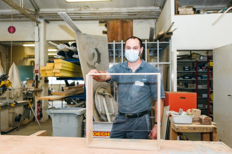 Un homme portant un uniforme bleu et un masque facial se tient debout dans un atelier. Il tient un cadre carré en bois. Différents outils, matériaux et équipements sont visibles autour de l'atelier, notamment des scies, des étagères et des établis, emblématiques du dévouement des Héros Méconnus du CH d'Angoulême.