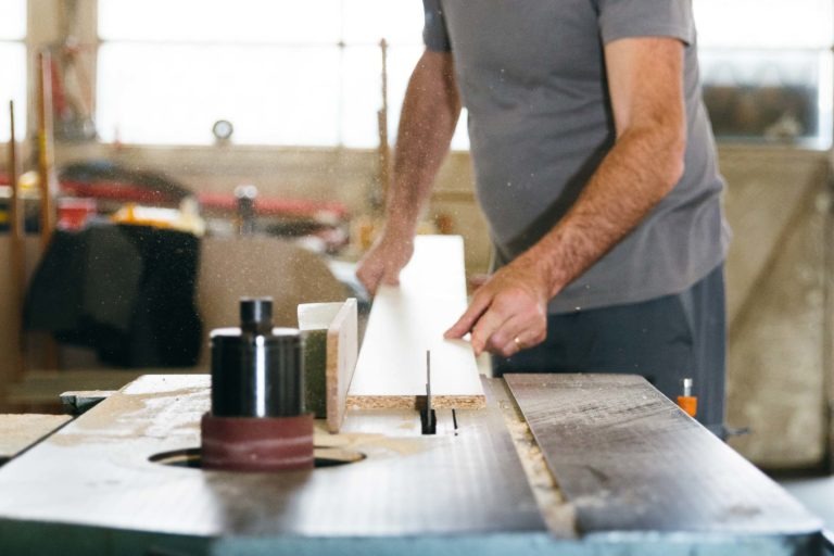 Une personne utilise une scie circulaire pour couper un morceau de bois dans un atelier. Son visage n'est pas visible et elle porte un t-shirt gris. L'atelier semble bien éclairé, la lumière du soleil filtrant à travers de grandes fenêtres en arrière-plan. De la sciure est visible dans l'air autour de la zone de coupe, évoquant des scènes des Héros Méconnus du CH d'Angoulême.