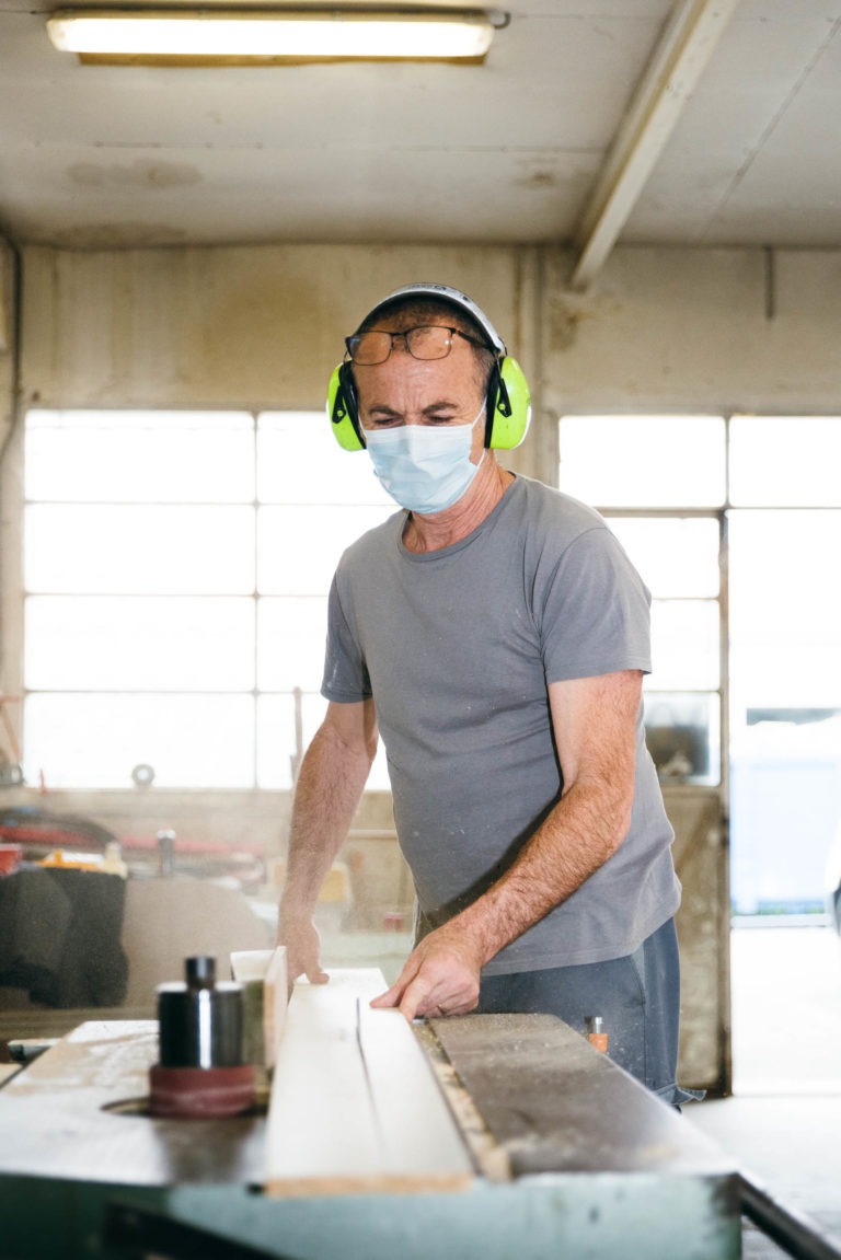 Un homme au crâne chauve et aux lunettes sur la tête se tient debout dans un atelier. Il porte un t-shirt gris, des cache-oreilles verts et un masque chirurgical bleu, et fait fonctionner une machine à bois. L'atelier est agrémenté de grandes fenêtres en arrière-plan et de divers outils autour de lui. Il est véritablement l'un des Héros Méconnus du CH d'Angoulême.