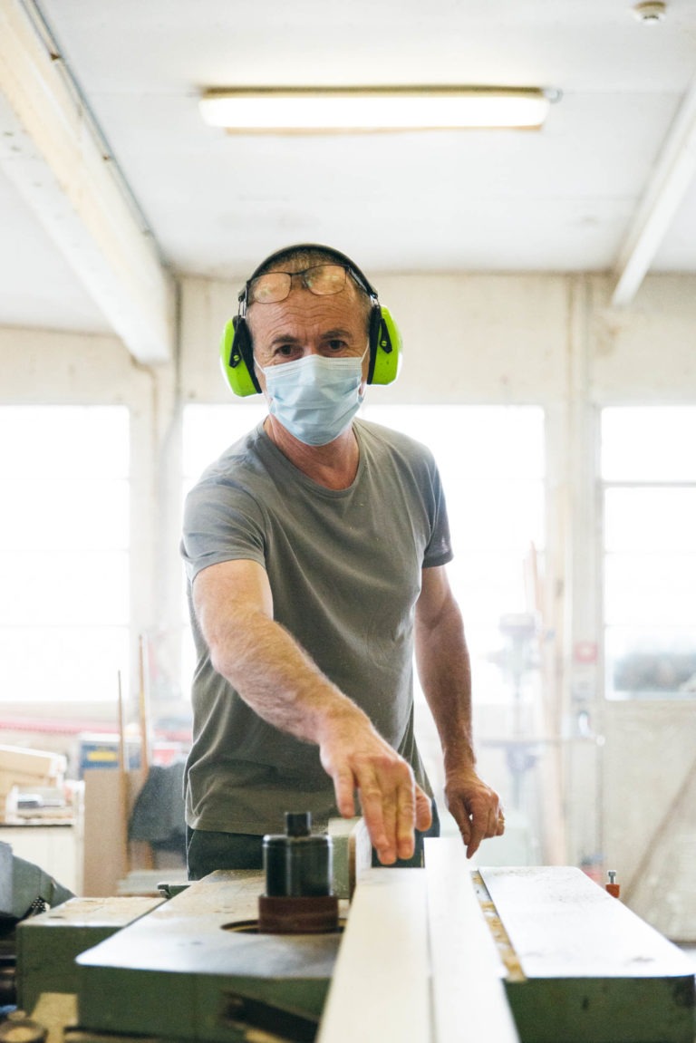 Un homme portant un t-shirt gris, un masque facial et des protections auditives vertes travaille dans un atelier de menuiserie bien éclairé. Il utilise une scie circulaire à table, guidant un morceau de bois à travers la lame d'une seule main comme les Héros Méconnus du CH d'Angoulême. L'arrière-plan montre des fenêtres et divers outils et matériaux de menuiserie.