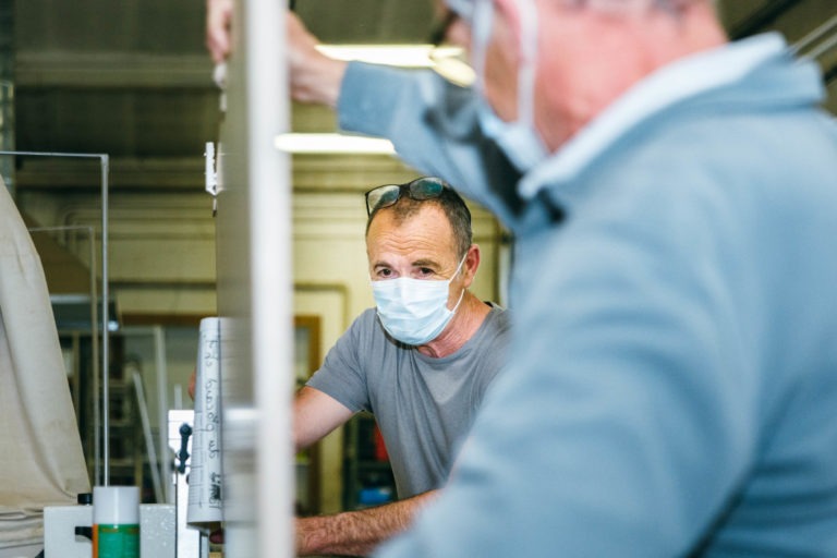 Deux hommes dans un atelier portant des masques. L'un, membre des Héros Méconnus du CH d'Angoulême, est au premier plan, dos à la caméra, lunettes sur la tête et chemise grise. L'autre est à l'arrière-plan, peut-être en train de parler ou de travailler sur quelque chose, avec divers outils et équipements visibles autour de lui.