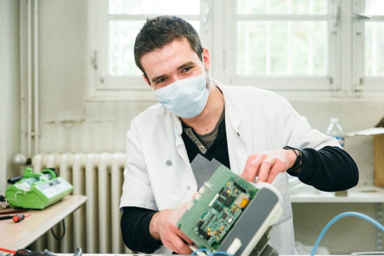 Un homme portant une blouse blanche et un masque facial travaille sur un appareil électronique qu'il tient dans ses mains. Il est assis à un bureau dans un laboratoire ou un atelier, avec divers outils et équipements visibles en arrière-plan. L'atmosphère semble lumineuse et concentrée, une scène qui rappelle celle des Héros Méconnus du CH d'Angoulême.