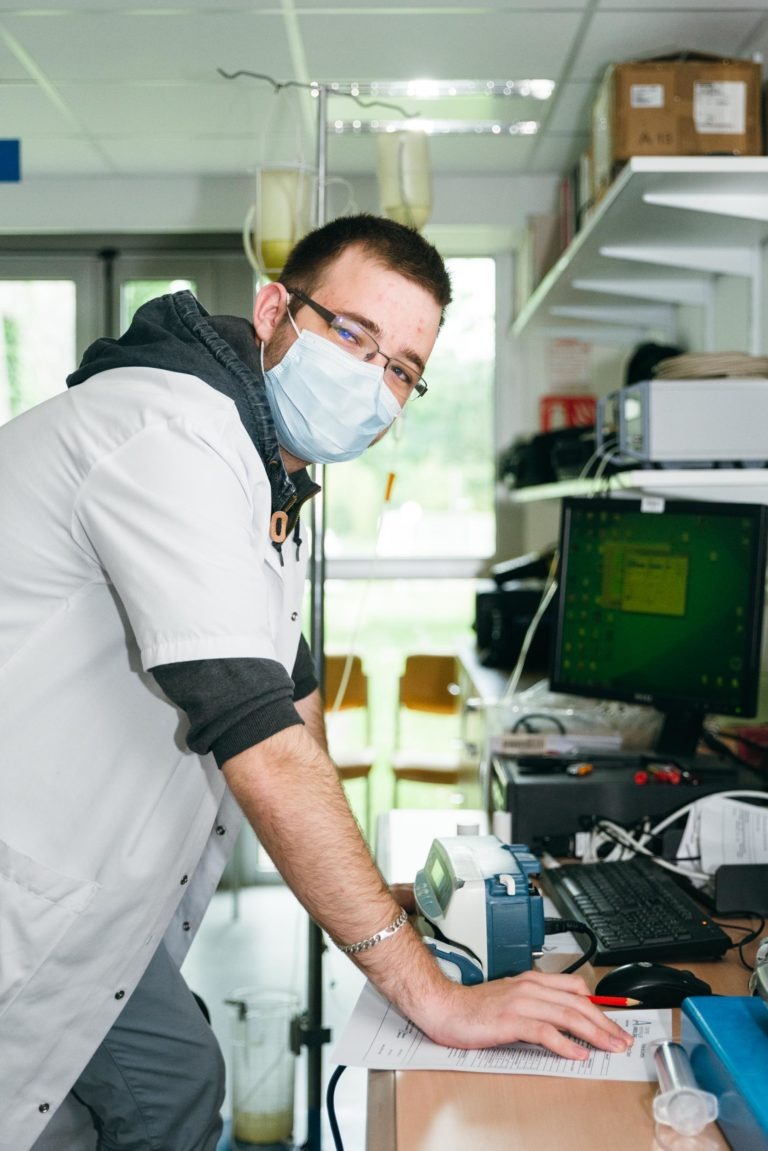 Une personne portant une blouse blanche et un masque bleu pose en s'appuyant sur un bureau dans un laboratoire. L'individu est entouré de divers équipements de laboratoire, dont un écran d'ordinateur et des instruments scientifiques. Des étagères avec des boîtes et des livres se trouvent sur le côté droit - de véritables Héros Méconnus du CH d'Angoulême.