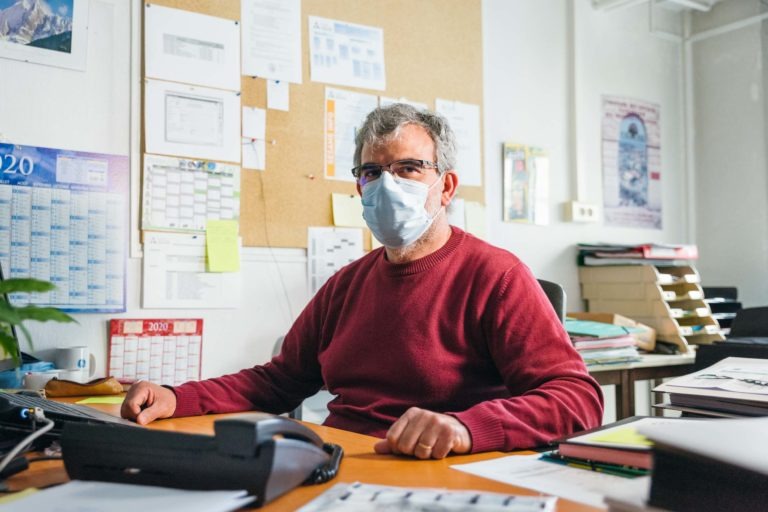 Un homme aux cheveux gris et aux lunettes, portant un pull rouge et un masque chirurgical bleu, est assis à un bureau encombré. Derrière lui se trouvent des tableaux en liège remplis de papiers et de calendriers des Héros Méconnus du CH d'Angoulême. Le bureau est doté d'un téléphone, de dossiers, de documents éparpillés et d'une pile de papiers à sa gauche.