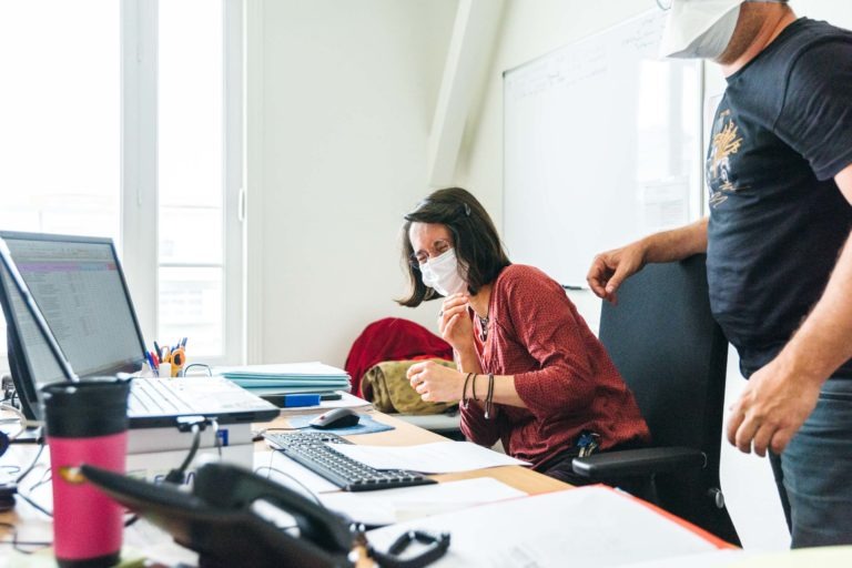 Deux personnes dans un bureau portent des masques. L'une, assise à un bureau avec plusieurs écrans d'ordinateur, rit tandis que l'autre, debout à côté d'elle, pointe l'écran du doigt. Le bureau est encombré de papiers, d'une tasse de voyage rose et de fournitures de bureau en arrière-plan - de véritables Héros Méconnus du CH d'Angoulême.