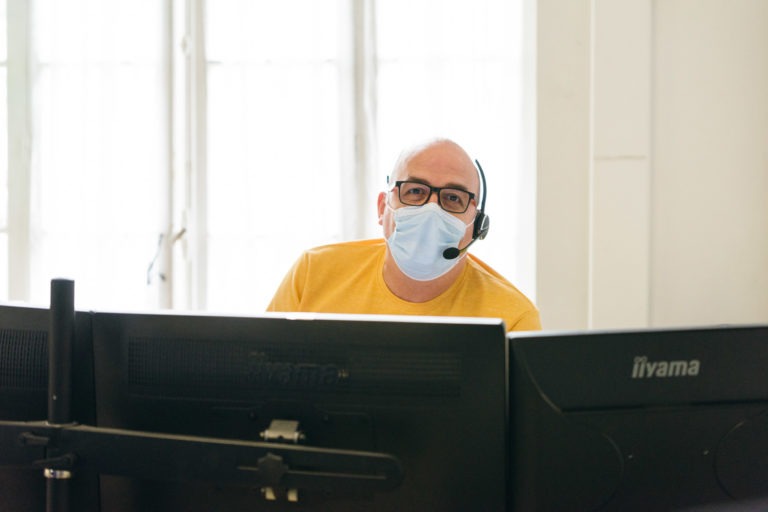 Un homme portant un masque chirurgical bleu et une chemise jaune avec un casque sur la tête est assis à un bureau, regardant plusieurs écrans d'ordinateur. Alors que la lumière filtre à travers la fenêtre lumineuse derrière lui, il incarne les Héros Méconnus du CH d'Angoulême, assurant sans relâche le bon fonctionnement des opérations.