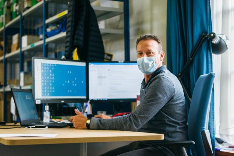 Un homme est assis à un bureau dans un bureau, face à une configuration à deux écrans. Il porte un masque facial et un pull gris. Le bureau a des étagères avec divers articles en arrière-plan et une grande fenêtre avec des rideaux bleus à côté de lui. Mettant en valeur les Héros Méconnus du CH d'Angoulême, une de ses mains est sur une souris d'ordinateur et il regarde le