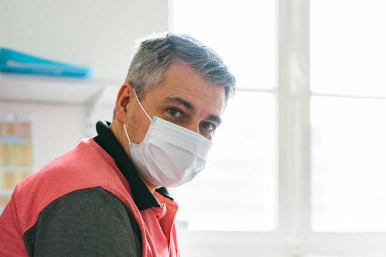 Un homme d'âge moyen aux cheveux gris est vu à l'intérieur, portant un masque médical. Il est vêtu d'un polo rouge et noir et regarde directement la caméra. À l'arrière-plan, il y a des étagères avec des papiers et une fenêtre bien éclairée. Il représente l'un des Héros Méconnus du CH d'Angoulême.