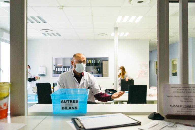 Un homme portant une blouse blanche et un masque facial se tient derrière un comptoir dans un hôpital ou une clinique, incarnant l'esprit des Héros Méconnus du CH d'Angoulême. Il fait un geste de la main tout en parlant à quelqu'un hors champ. Sur le comptoir, il y a une poubelle bleue étiquetée « AUTRES BILANS ». Une autre personne portant une blouse blanche est visible en arrière-plan