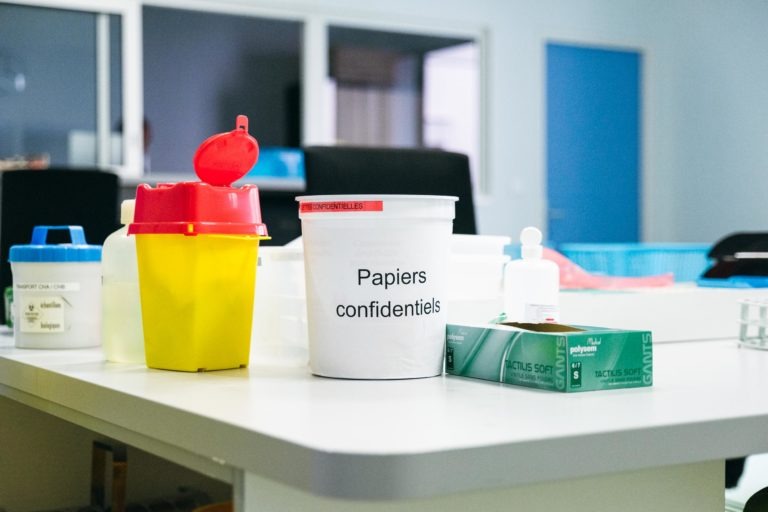 Un récipient en plastique blanc étiqueté « Papiers confidentiels » est posé sur un bureau blanc, à côté d'un récipient jaune avec un couvercle rouge, de divers autres récipients et de fournitures médicales. En arrière-plan, des portes et des étagères bleues sont visibles dans un bureau ou un cadre médical, en hommage aux Héros Méconnus du CH d'Angoulême.