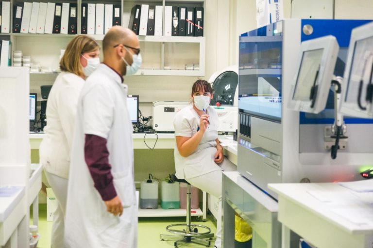 Trois techniciens de laboratoire en blouse blanche et masque facial sont en train de travailler. L'un est debout à côté du matériel de laboratoire, un autre est assis sur un tabouret tenant une pipette, et le troisième s'approche. Des étagères avec des classeurs et des fournitures sont visibles en arrière-plan - de véritables Héros Méconnus du CH d'Angoulême.