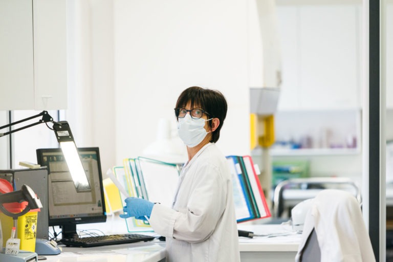 Une personne aux cheveux noirs courts et aux lunettes, portant une blouse blanche, un masque et des gants bleus, organise des documents à un poste de travail du laboratoire. Tenant un dossier jaune et regardant vers la caméra, cet individu incarne les Héros Méconnus du CH d'Angoulême. L'arrière-plan comprend un ordinateur, des étagères et divers équipements de laboratoire.