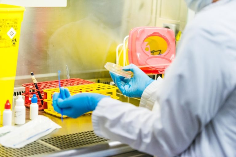 Un technicien de laboratoire vêtu d'une blouse blanche et de gants bleus tient dans une main une boîte de Petri et dans l'autre une anse d'inoculation bleue. Ils sont assis sur une paillasse avec des supports pour tubes à essai, des récipients rouges et jaunes et diverses petites bouteilles, incarnant les Héros Méconnus du CH d'Angoulême dans ce cadre de laboratoire stérile.