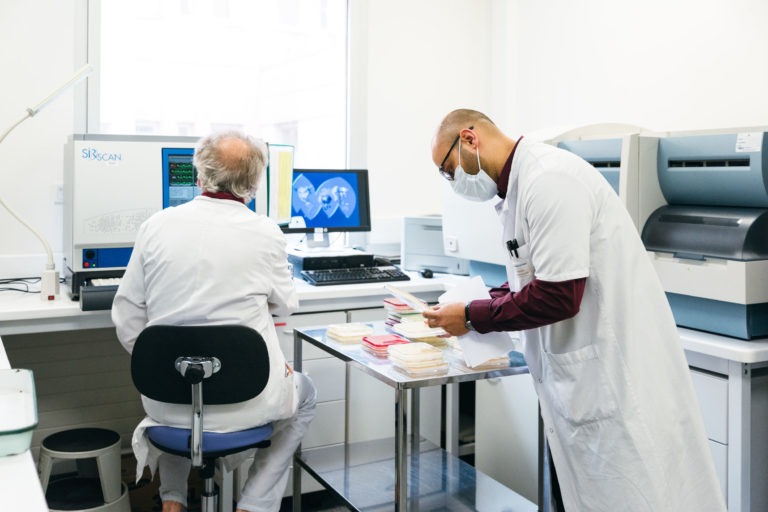 Deux scientifiques en blouse blanche travaillent dans un laboratoire lumineux et propre. L'un est assis devant un ordinateur avec des images médicales sur les écrans, tandis que l'autre est debout, organisant des documents sur une table. Tous deux sont concentrés sur leurs tâches, incarnant l'esprit des Héros Méconnus du CH d'Angoulême.