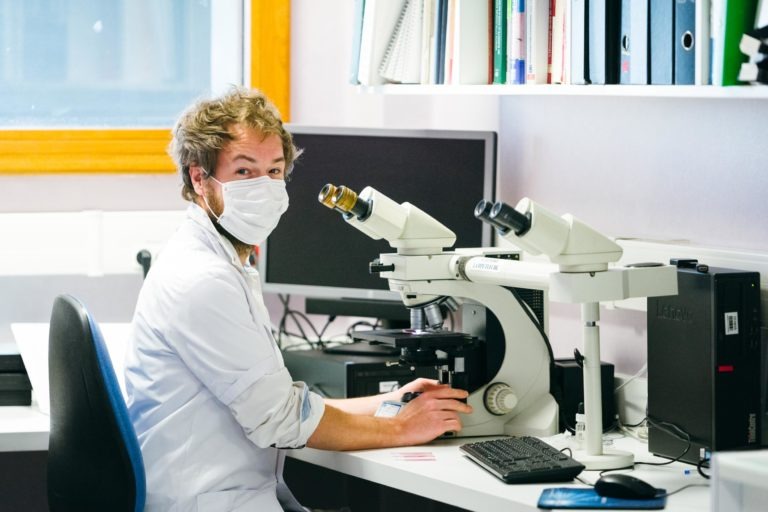 Une personne aux cheveux bouclés, portant une blouse blanche et un masque facial, est assise à un bureau en train d'utiliser un microscope dans un laboratoire. Des livres et des dossiers sont soigneusement disposés sur une étagère au-dessus du bureau. Un écran d'ordinateur est en arrière-plan. L'un des Héros Méconnus du CH d'Angoulême, ils semblent regarder la caméra tout en travaillant.