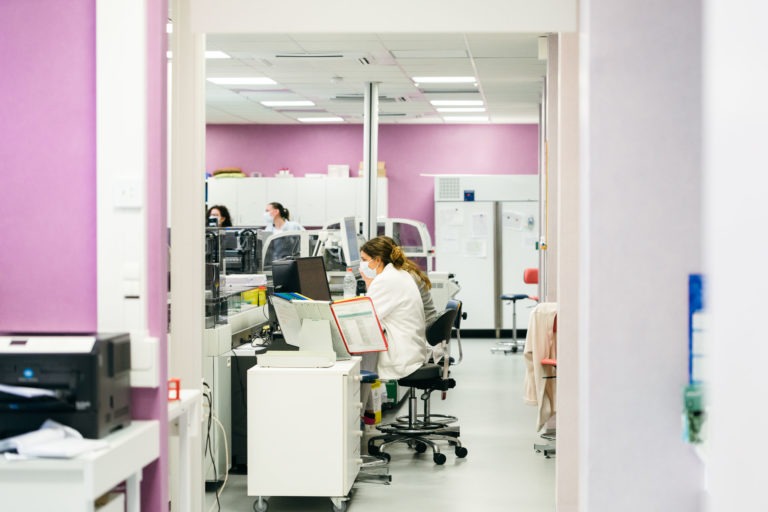 Une personne portant une blouse blanche et un masque est assise à un bureau d'ordinateur dans un laboratoire lumineux aux murs violets. D'autres membres du personnel et équipements du laboratoire sont visibles à l'arrière-plan. Le laboratoire spacieux et bien éclairé comporte divers postes de travail, des armoires de rangement et un cadre de porte au premier plan, illustrant les Héros Méconnus du CH d'Angoulême.