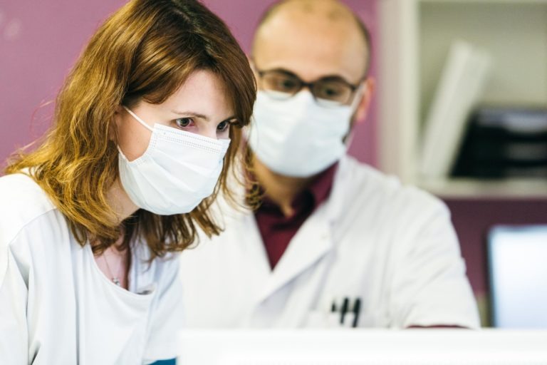 Deux professionnels de santé, une femme et un homme, portent des blouses blanches et des masques de protection. La femme au premier plan est concentrée sur un écran d'ordinateur, tandis que l'homme à l'arrière-plan regarde également l'écran. Ils représentent les Héros Méconnus du CH d'Angoulême dans leur cadre clinique ou de laboratoire avec un mur violet.