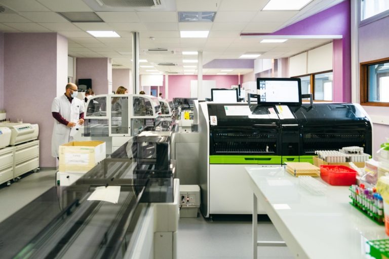 Un laboratoire spacieux avec des équipements et des postes de travail de pointe. Deux techniciens de laboratoire en blouse blanche et masque facial, les Héros Méconnus du CH d'Angoulême, se tiennent sur le côté gauche près de quelques machines. Le laboratoire dispose de grands analyseurs, de plateaux avec des tubes à essai et de diverses fournitures de laboratoire soigneusement organisées sur des tables.