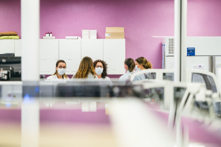 Un groupe de six individus, tous vêtus de blouses blanches et de masques faciaux, sont réunis pour discuter dans un laboratoire au mur rose vif. Des armoires et des boîtes blanches sont visibles à l'arrière-plan. La scène est observée à travers une vitre ou un équipement, ce qui donne un premier plan légèrement flou. Ce sont vraiment les Héros Méconnus du CH d'Angoulême.