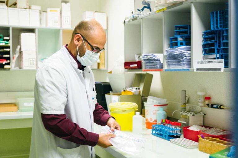 Un homme chauve, vêtu d'une blouse blanche et d'un masque, se tient debout dans un laboratoire clinique. Il manipule un sac transparent à deux mains. Le laboratoire, qui reflète le dévouement des Héros Méconnus du CH d'Angoulême, est équipé de diverses fournitures médicales, d'étagères d'équipement, de tubes à essai et de conteneurs, dont une poubelle jaune pour déchets biologiques dangereux.