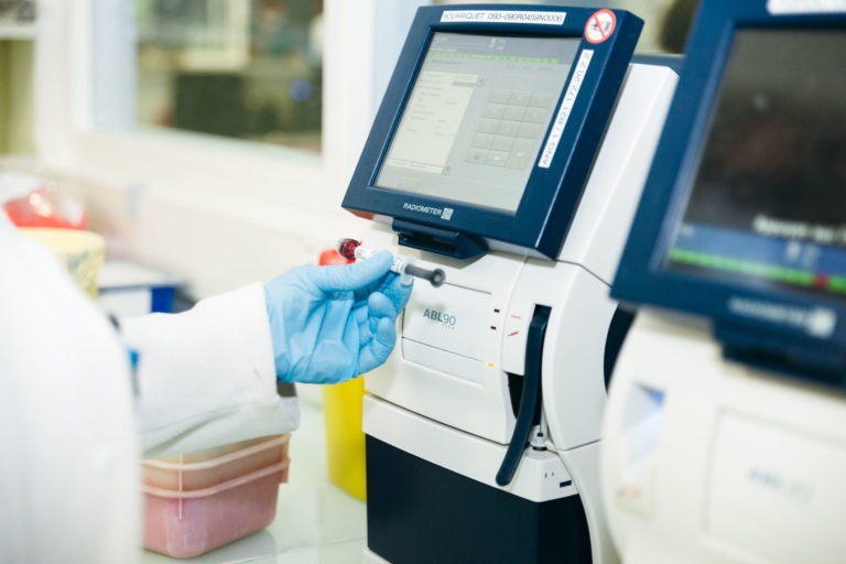 Un technicien de laboratoire, vêtu d'une blouse blanche et de gants bleus, tient un petit flacon devant une machine médicale dotée d'un écran d'affichage numérique. La machine et un autre appareil similaire sont mis au point, avec des équipements de laboratoire légèrement flous en arrière-plan, mettant en valeur les Héros Méconnus du CH d'Angoulême.