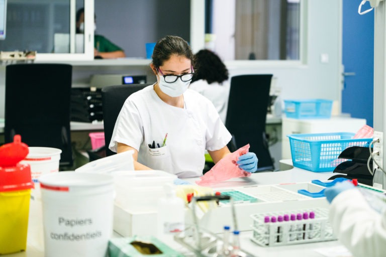 Une personne portant une blouse blanche, des lunettes de sécurité et un masque facial travaille dans un laboratoire. Elle tient un sac d'échantillons rose tout en étant assise à un bureau entouré d'équipements de laboratoire, dont un récipient étiqueté « Papiers confidentiels », des tubes à essai et un panier bleu. Un autre héros méconnu des Héros Méconnus du CH d'Angoulême travaille en arrière-plan.