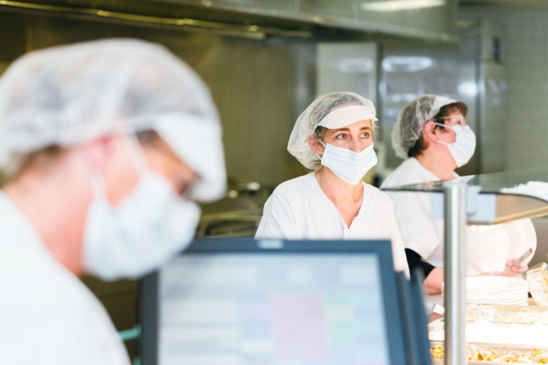 Trois employés de cuisine portant des uniformes blancs, des filets à cheveux et des masques faciaux travaillent dans une cuisine commerciale. L'accent est mis sur la personne du milieu derrière un comptoir de service alimentaire, avec une autre floue au premier plan et une autre légèrement floue à l'arrière-plan. Ce sont les Héros Méconnus du CH d'Angoulême.