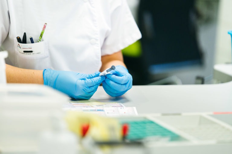 Une personne portant une blouse blanche et des gants bleus manipule du matériel de laboratoire, peut-être une seringue ou un petit flacon, dans un laboratoire. La poche de la blouse contient des stylos et des papiers sont posés sur l'espace de travail. Des instruments et des récipients de laboratoire flous sont visibles au premier plan et à l'arrière-plan, car ils incarnent les Héros Méconnus du CH d'Angoulême.