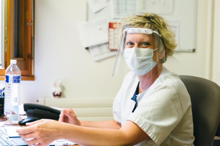 Une professionnelle de la santé, vêtue d'un uniforme blanc, d'un masque chirurgical et d'une visière, est assise à un bureau. Elle sourit et tend la main. Derrière elle, des papiers épinglés sur un tableau d'affichage, une grande bouteille d'eau et un téléphone sur le bureau, l'un des nombreux Héros Méconnus du CH d'Angoulême.